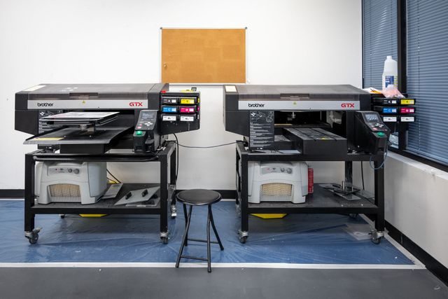 Two Brother GTX direct-to-garment printers on rolling carts side by side, with a stool between them