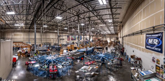 Panoramic view of the screen printing floor with two carousel presses, drying tables and an M&R banner on the wall