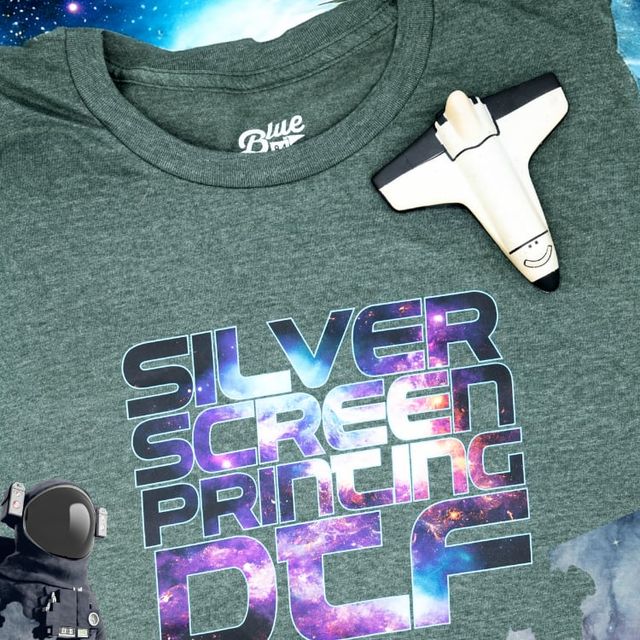 Heather green T-shirt with a galaxy-pattern DTF print reading Silver Screen Printing DTF, beside a toy space shuttle