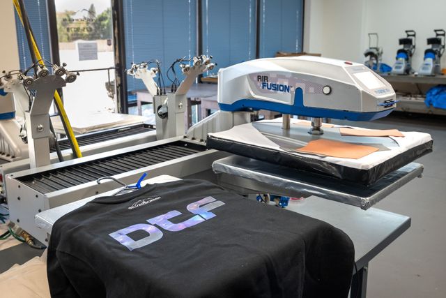 Hotronix Air Fusion heat press next to a black T-shirt with a DTF print, with shirt-loading stations and a conveyor dryer behind