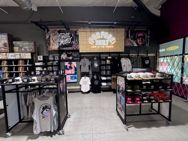 Retail shop with racks of printed t-shirts and hats under a lit Purpose Built, Home of the Trades sign
