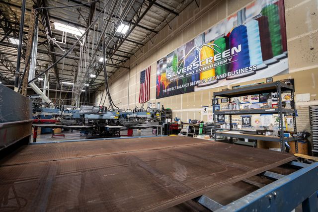Silverscreen screen printing floor with a conveyor dryer, automatic presses, an American flag and the Silverscreen Printing and Embroidery banner