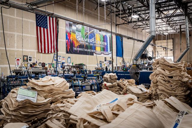 Silverscreen production floor stacked with tan tote bags, with an American flag and the Silverscreen banner on the wall