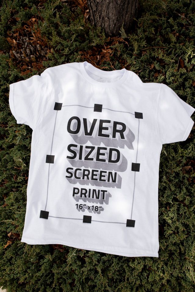 White T-shirt on a juniper shrub printed with a large 16 by 18 inch design reading Oversized Screen Print