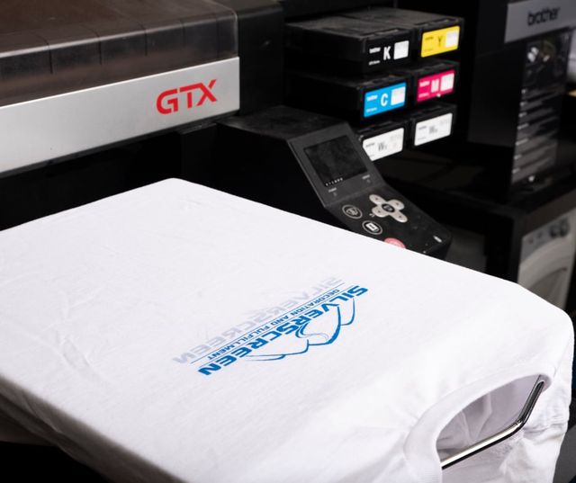 White T-shirt printed with the blue Silverscreen logo on the platen of a Brother GTX printer