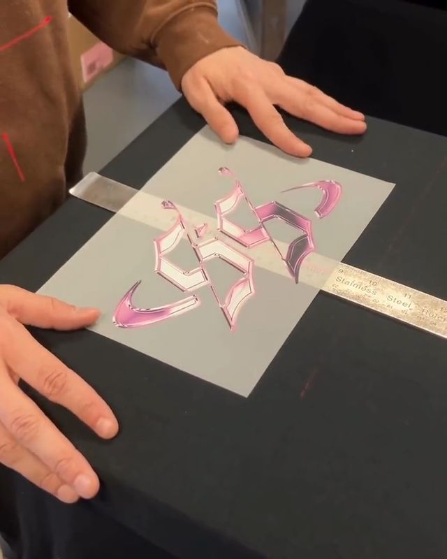 Hands smoothing a DTF transfer film printed with a pink chrome-effect gothic lettering design onto a black work surface, a steel ruler laid across it