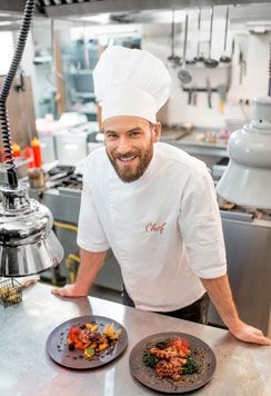 Smiling chef in a white jacket embroidered with the word Chef, standing behind two plated dishes in a restaurant kitchen