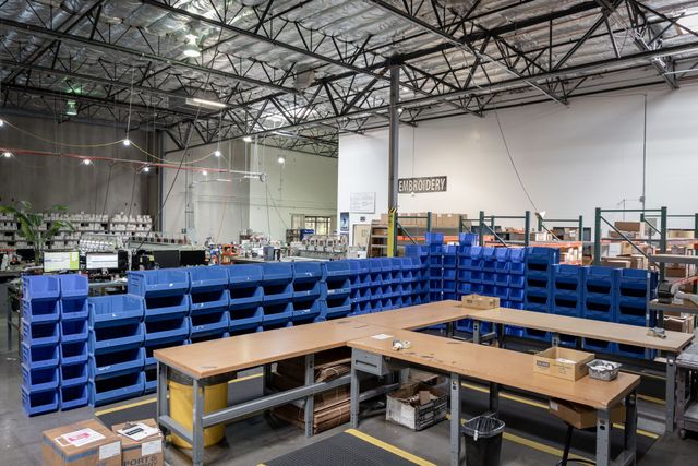 Fulfillment floor with walls of blue parts bins, packing tables and an Embroidery sign in the back