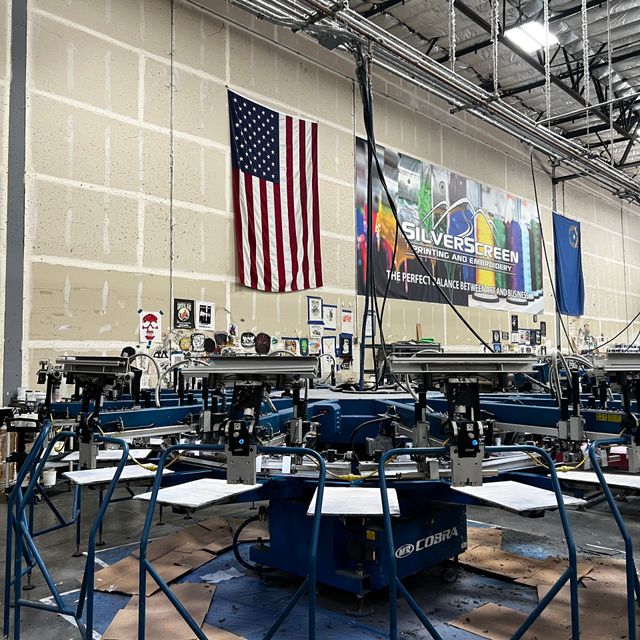 Automatic screen printing press on the Silverscreen production floor, with an American flag, a Nevada flag and a Silverscreen banner on the wall