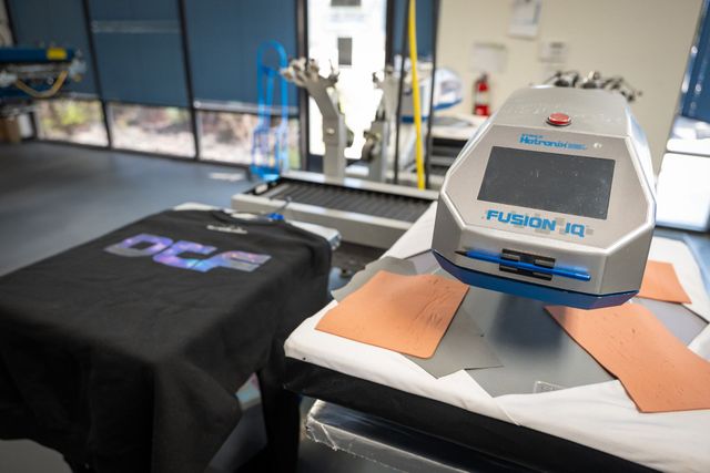 Black T-shirt with a holographic DTF print beside a Hotronix Fusion IQ heat press, with transfer film on the press platen